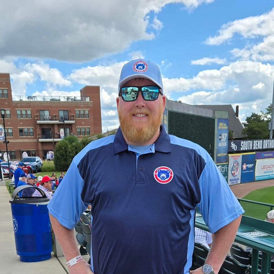 A man wearing a navy blue and light blue Chicago Cubs polo shirt, gray shorts, sunglasses, and a Cubs cap, standing at a baseball stadium.
